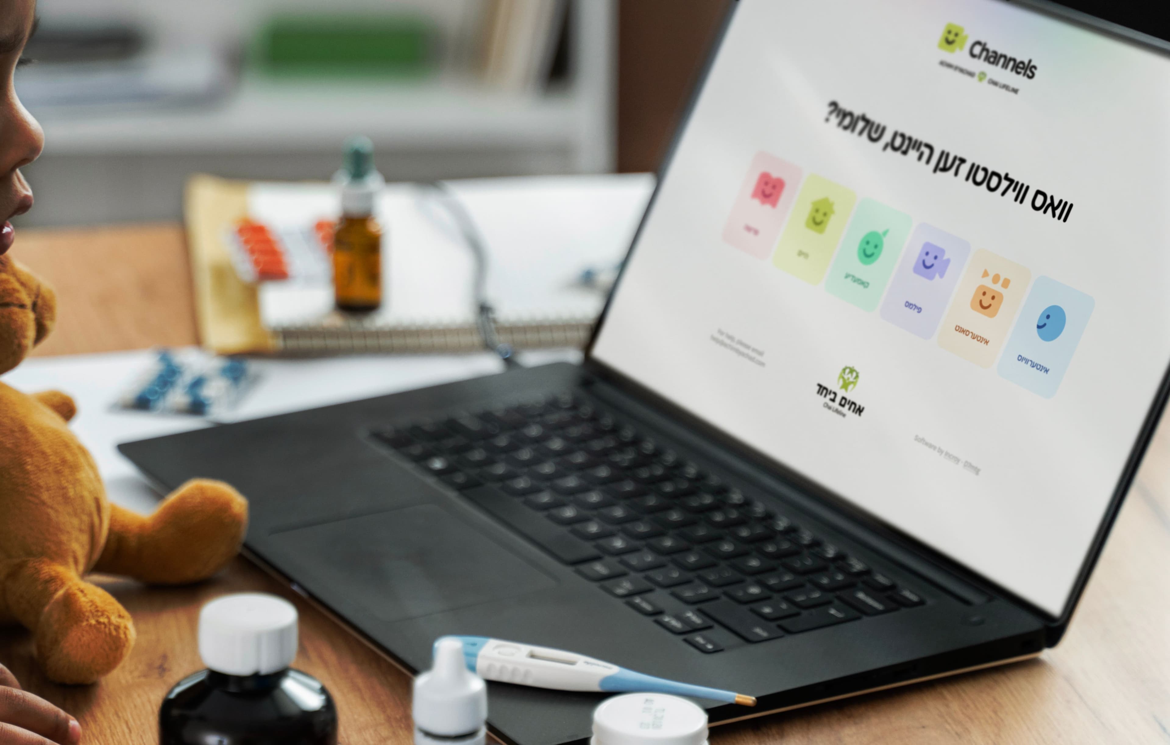 Laptop displaying Channels app with colorful smiley-faced buttons in Hebrew, with a stuffed toy nearby in a hospital setting